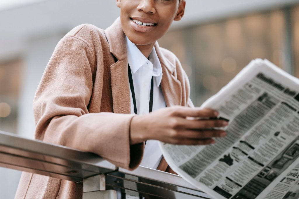 Crop happy African American businesswoman in formal clothes reading newspaper and looking away with smile while leaning on ra