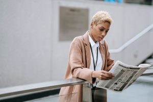 Serious young African American businesswoman in formal clothes leaning on business center building railing and reading articl