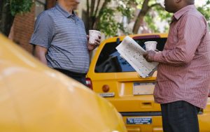 Crop anonymous multiracial taxi drivers with takeaway coffee and newspaper speaking near yellow cars in town — Die besten Bon